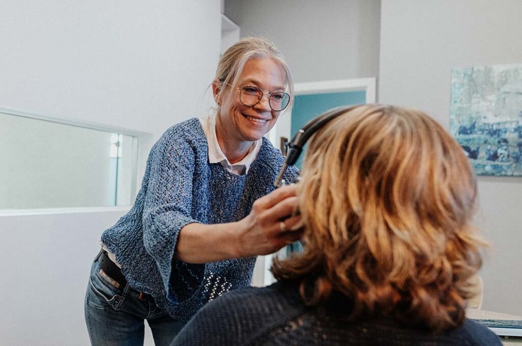 Eine lächelnde Frau mit Brille hilft einer Person mit hellbraunen Haaren beim Aufsetzen von Kopfhörern